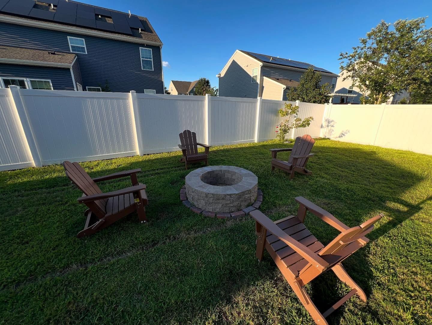 Backyard with a stone fire pit surrounded by four wooden chairs on a grassy lawn, bordered by white fencing