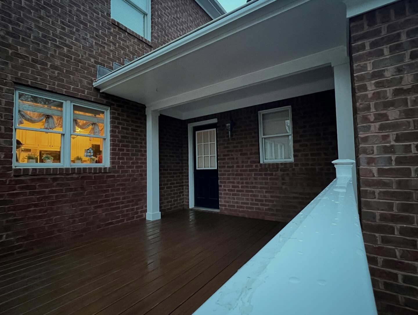 Covered brick apartment patio with dark wooden deck, white railing, and a lit window at dusk
