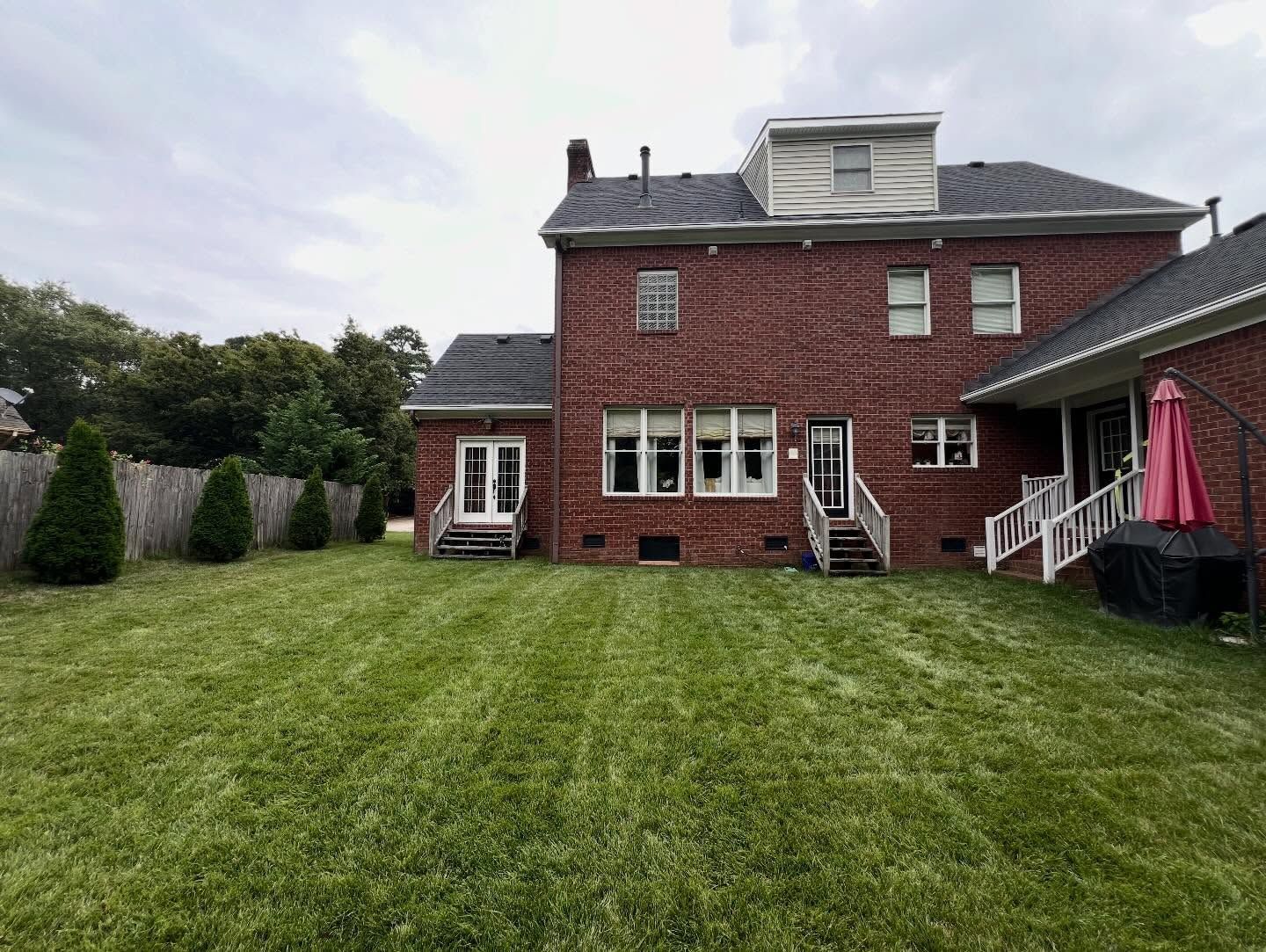 Backyard lawn facing a red brick house with patio chairs, windows, and a pink umbrella