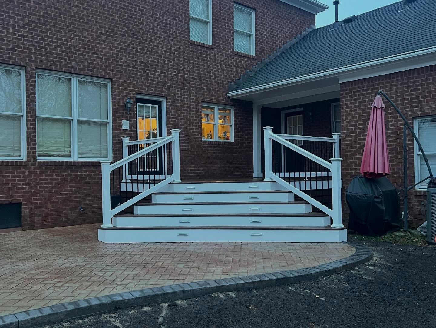Brick house patio with white railing, steps, and a pink umbrella beside a lit window