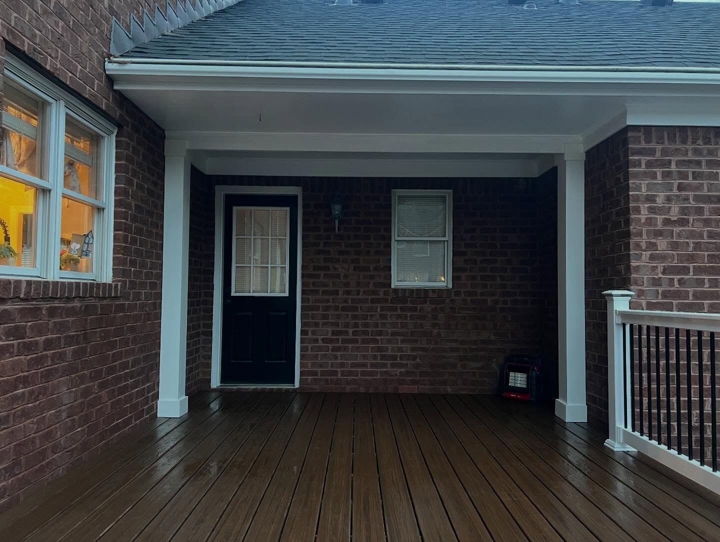 Covered wooden porch with brick walls, a black door, and wet floor after rain.