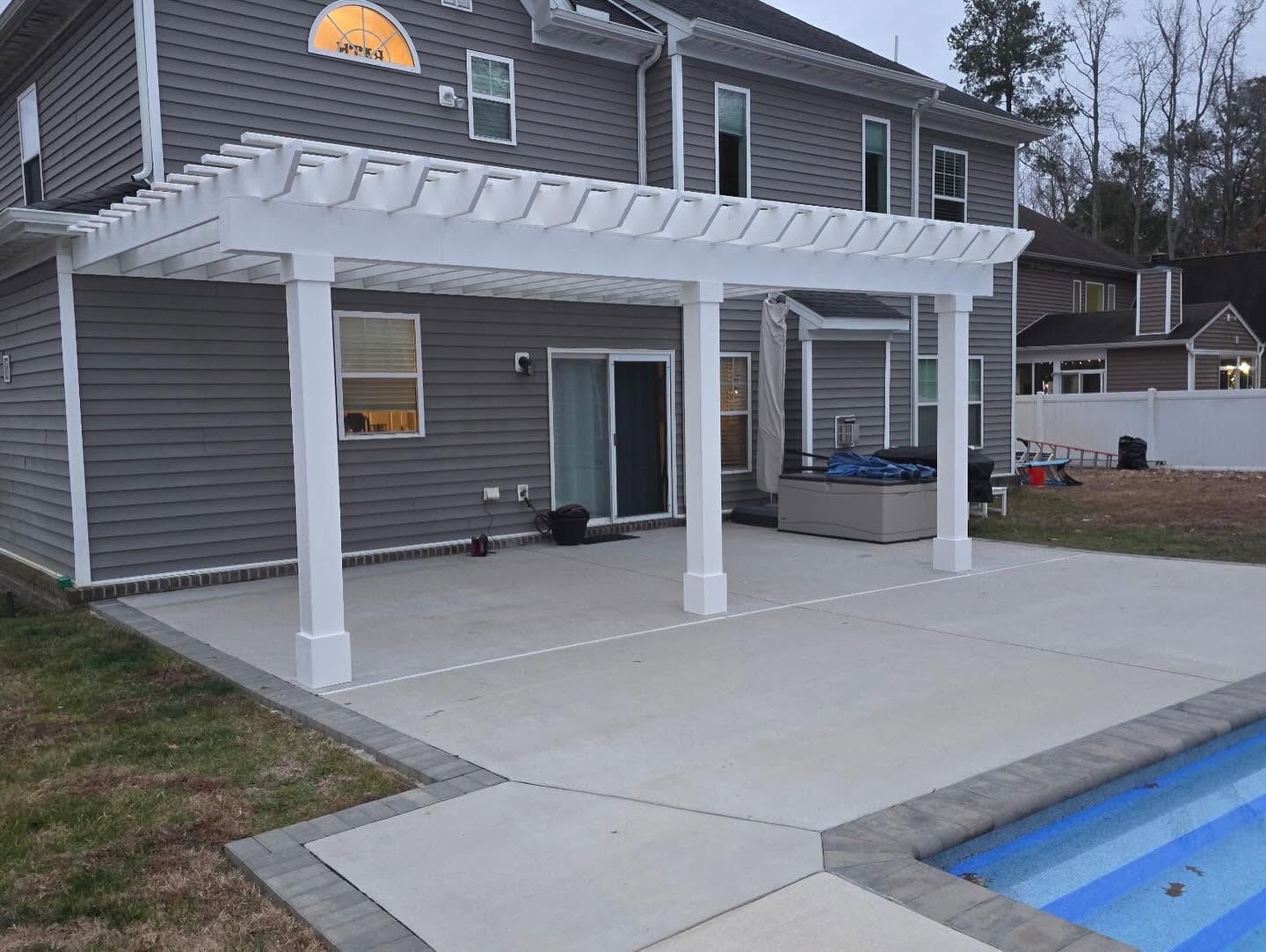 Backyard patio with white pergola and concrete slab beside a house, next to a pool