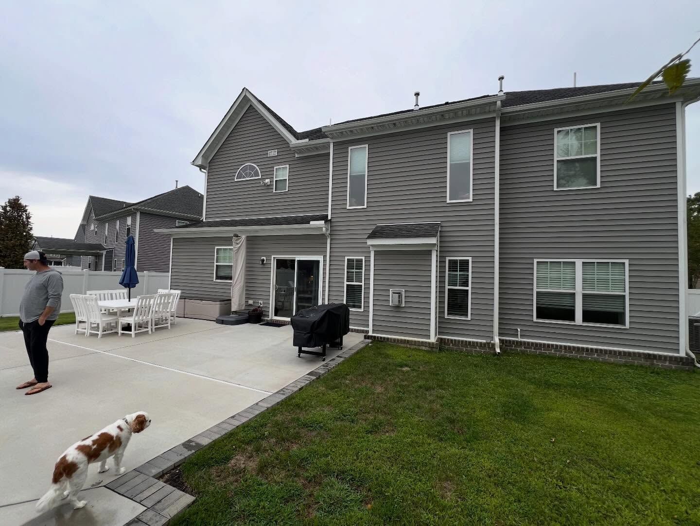 Backyard of gray two-story house with patio, lawn, grill, and two dogs near a person by the driveway