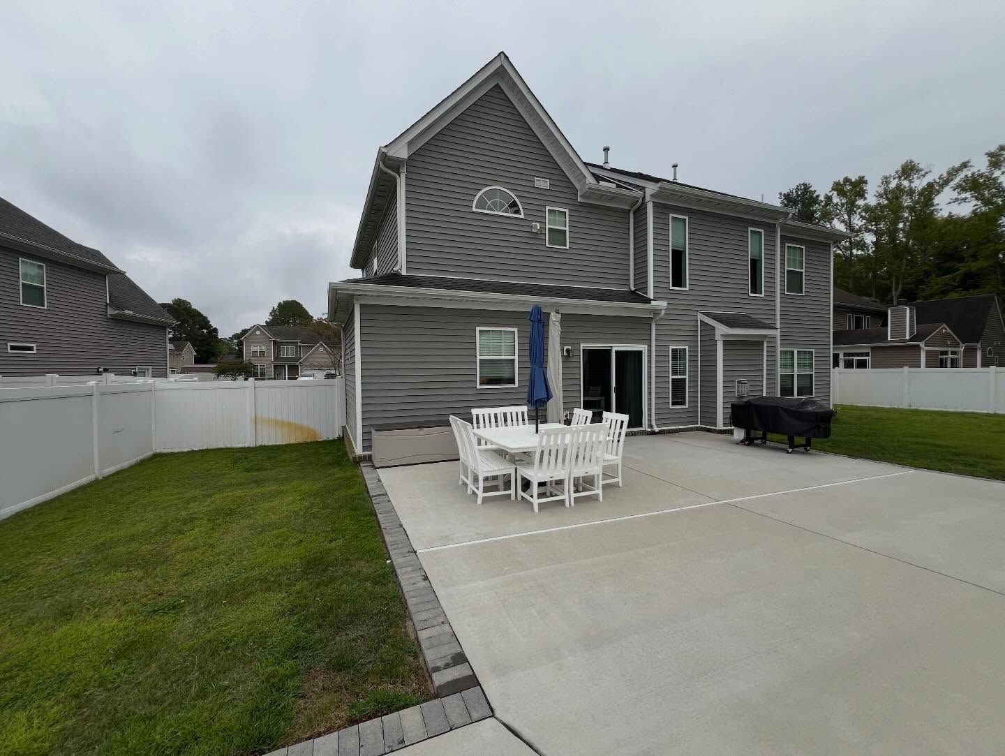 Backyard of gray house with white fence, patio table, and large concrete driveway under cloudy sky