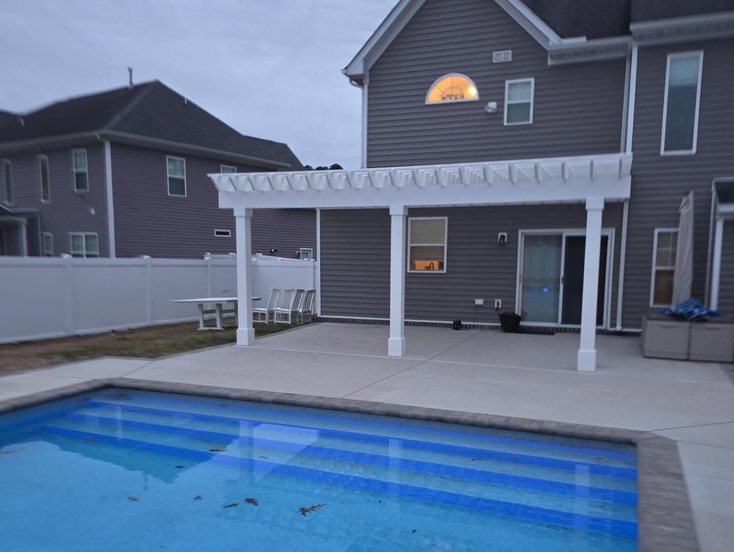 Backyard patio with a covered deck, swimming pool, and a two-story house at dusk
