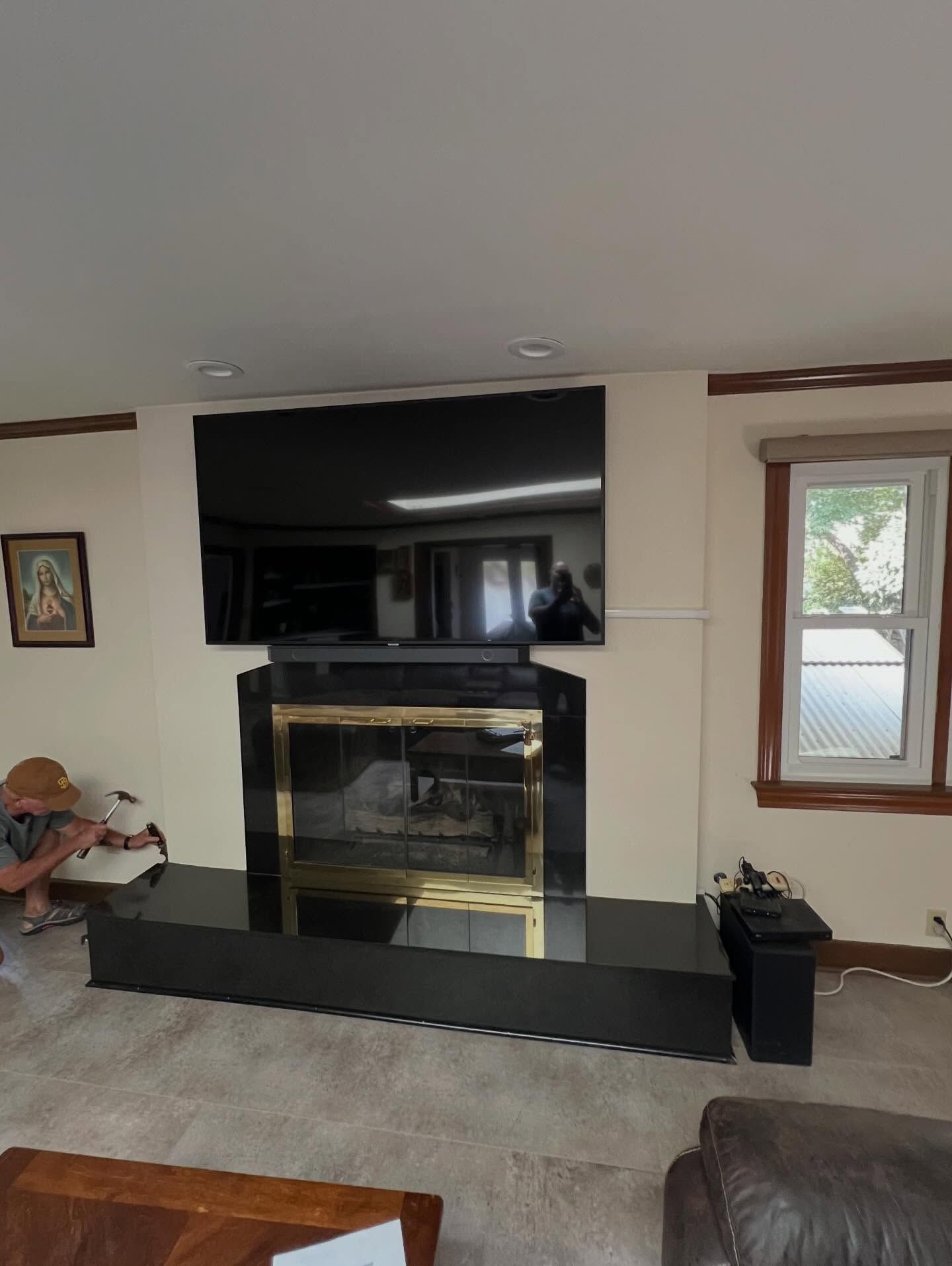 Living room with a black fireplace, wall-mounted TV, and windows; person crouches near the left wall