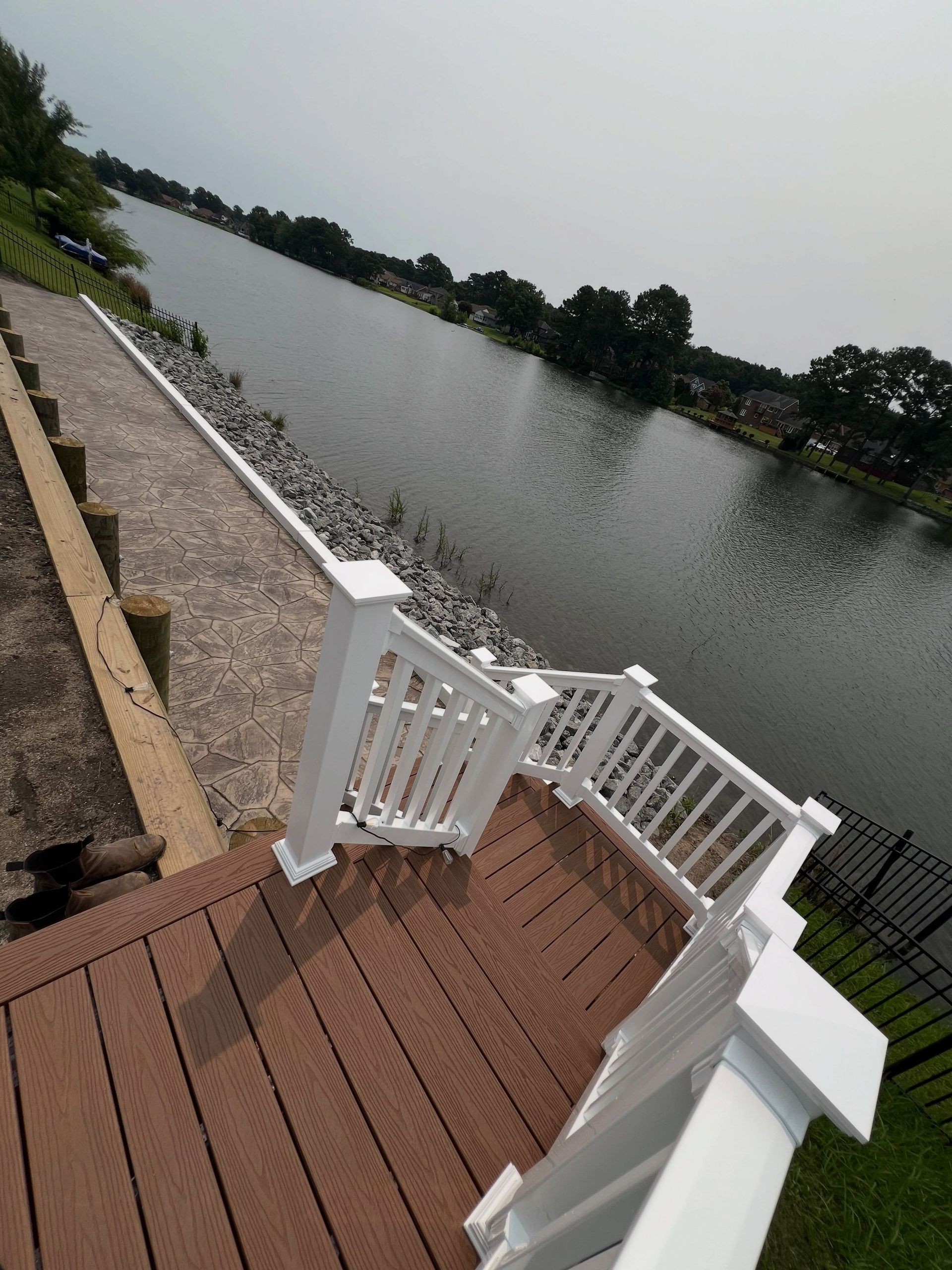 Wooden deck stairs with white railings descending to a calm lakeside walkway under an overcast sky