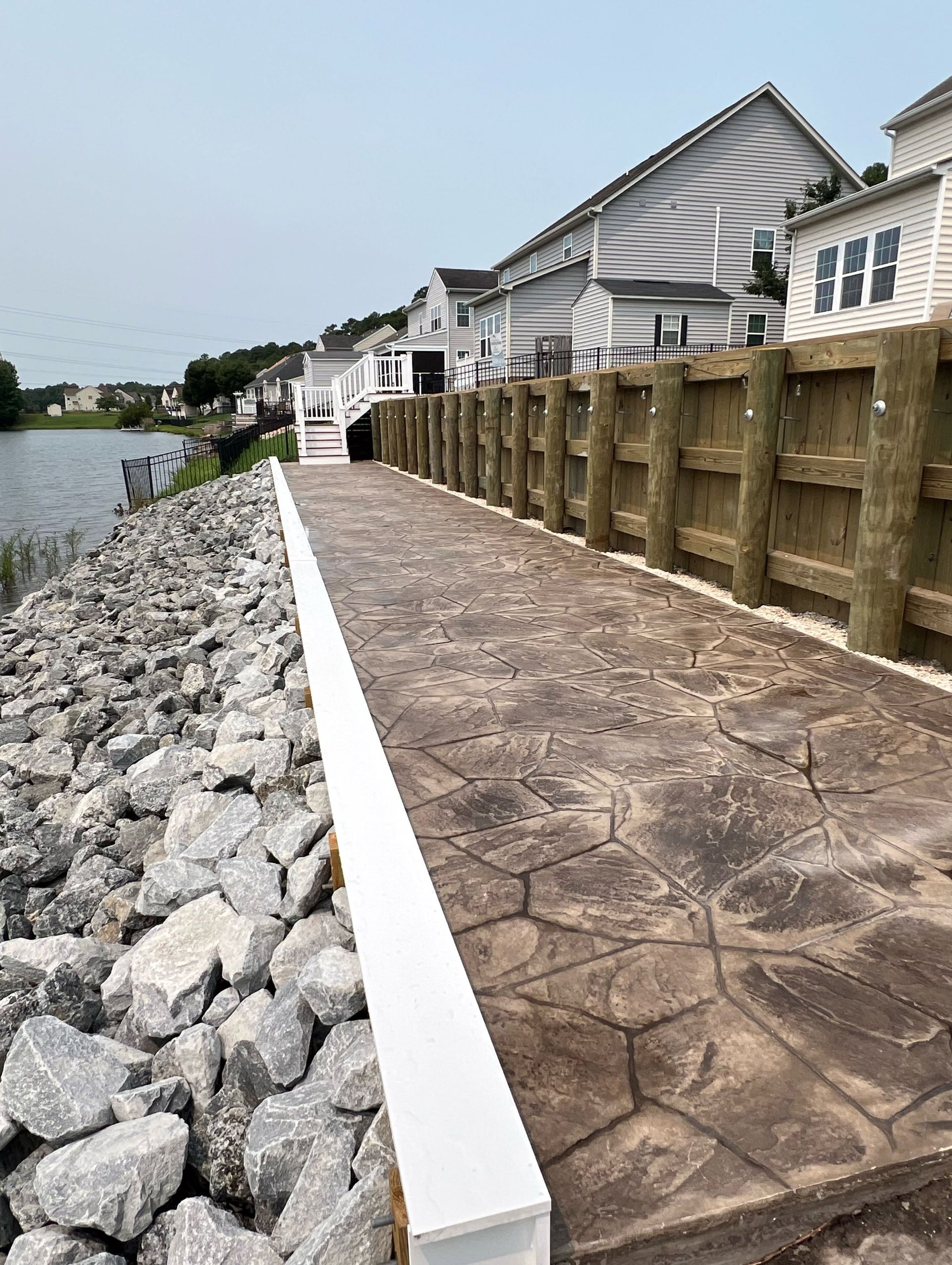 Rock-lined waterfront walkway beside houses and a wooden fence along the shore