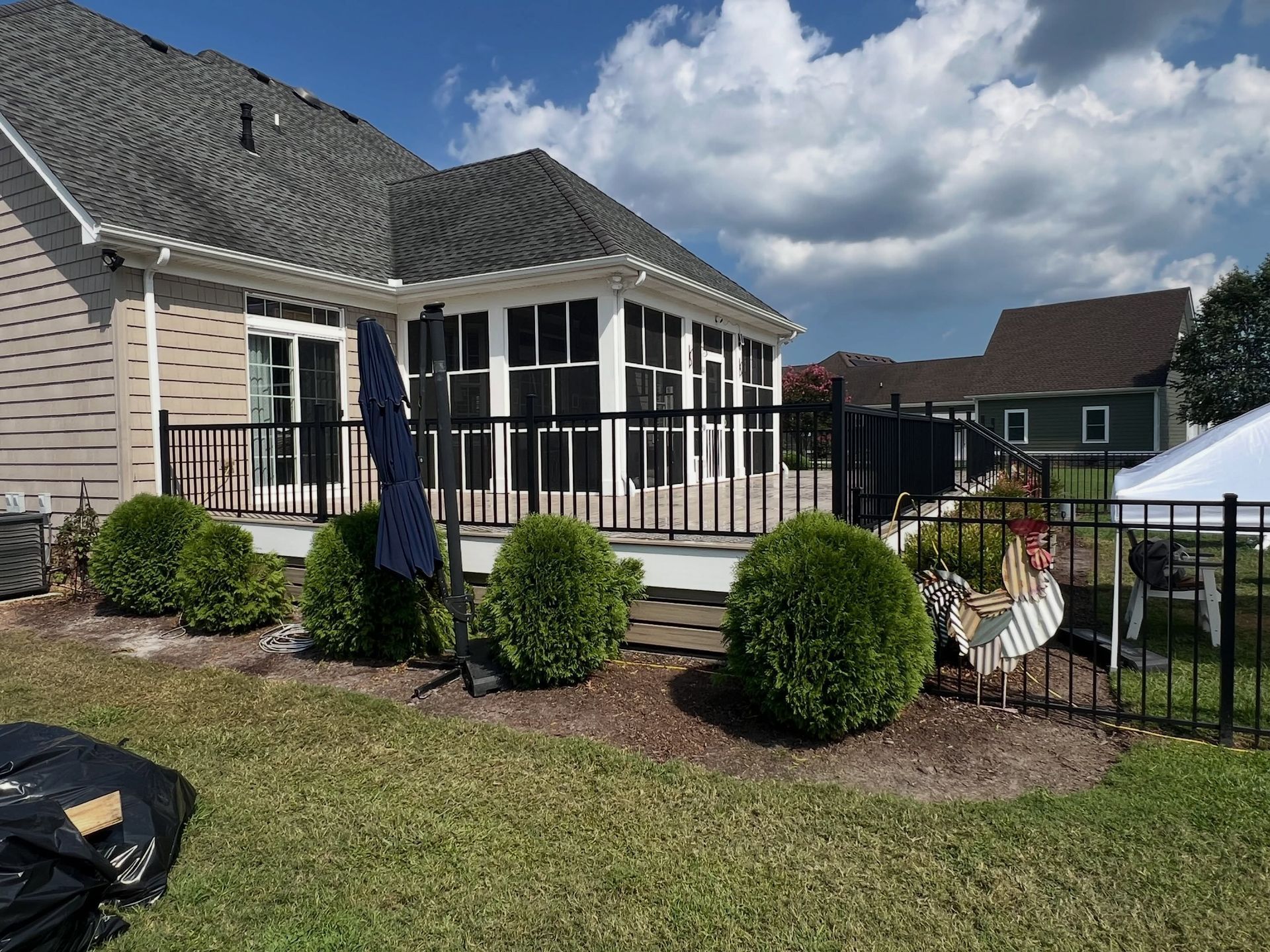 Backyard patio with screened porch, black fence, green lawn, and shrubs under a cloudy sky