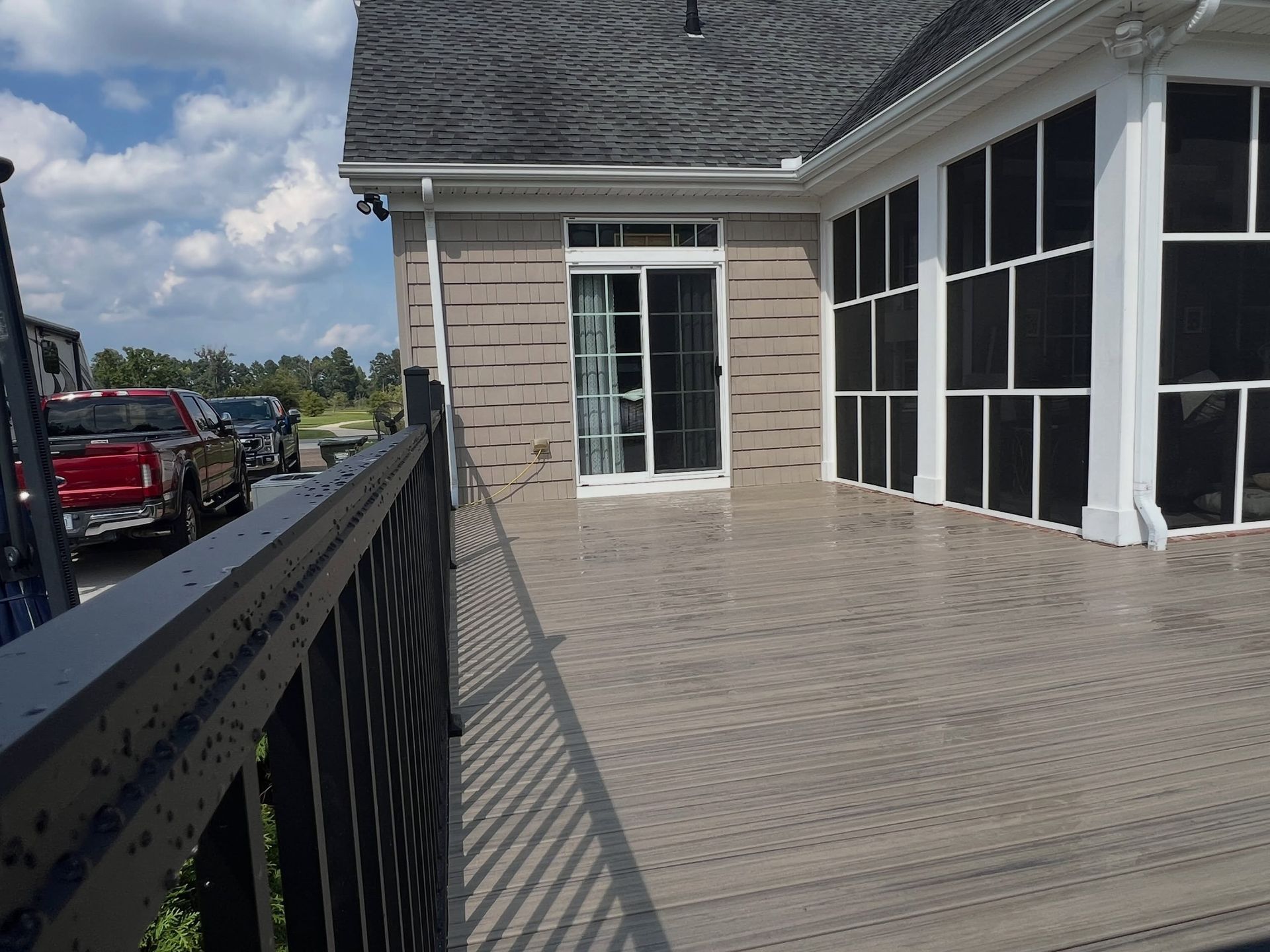 Covered outdoor patio with gray patterned flooring, white-framed windows, and a black railing beside a parking lot