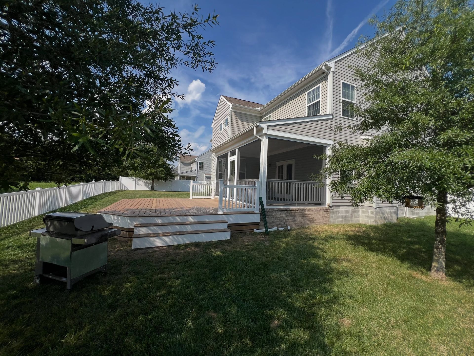 Backyard with wooden deck, white fence, grill, and two-story house under a blue sky