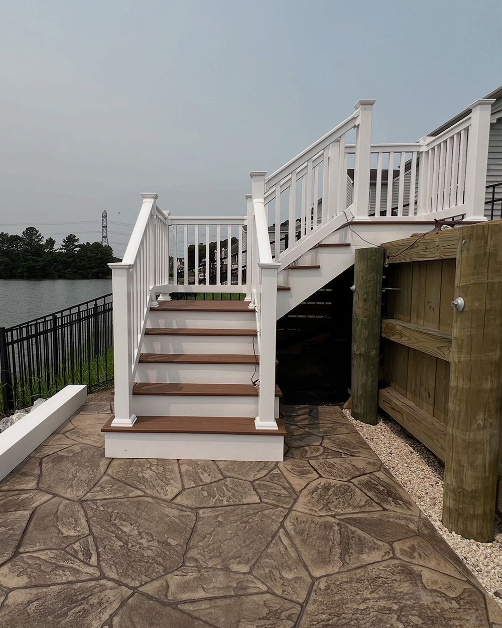 White wooden stairs and deck railing beside a stone patio near water, with a wooden fence on the right.