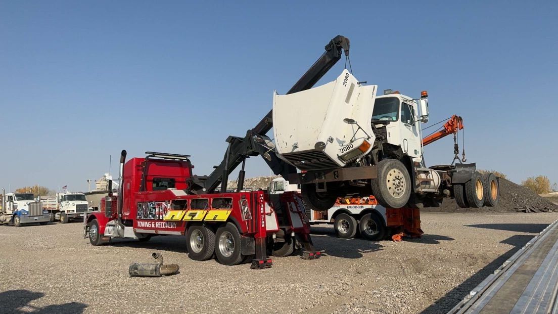 A red tow truck is towing a white truck in a gravel lot.