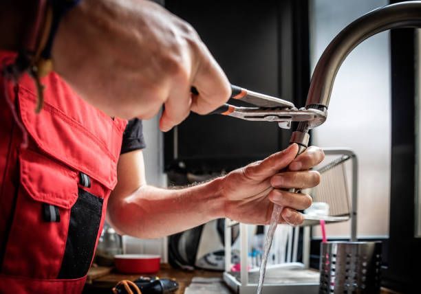 Plumber in red overalls uses pliers to repair a leaky kitchen faucet; water streams.