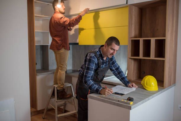Two men installing yellow kitchen cabinets; one on a ladder, other looking at plans.