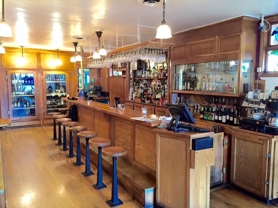 A wooden bar with stools, bottles, and glasses. Lights hang above the bar.