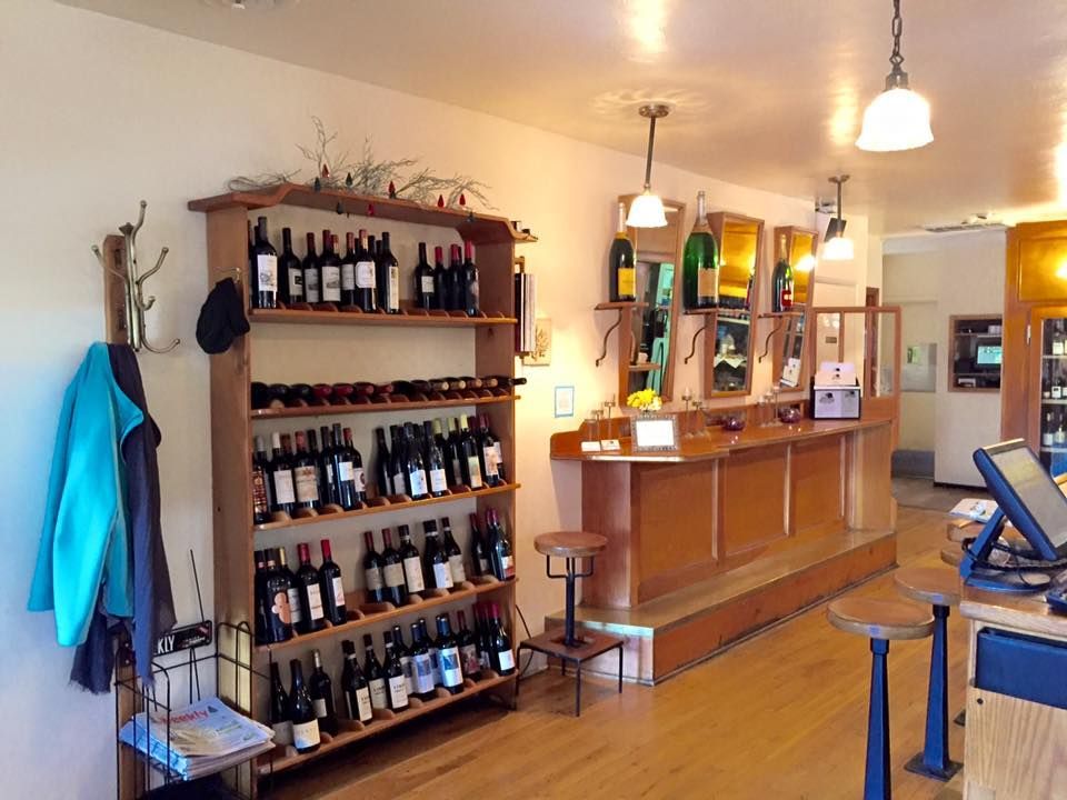 Wine shop interior, wooden shelves with bottles, a bar with stools, and hanging coats.