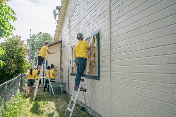 People in yellow shirts painting a light-colored house exterior. They are on ladders and covering windows. Sunny day.