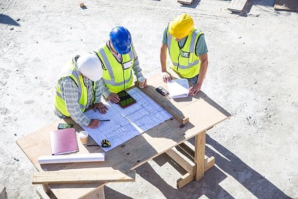 Construction workers in hard hats reviewing blueprints at a makeshift table on a sunny construction site.