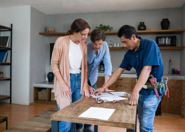 A plumber reviewing plans with a couple at a wooden table in a modern home.