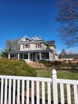 Two-story beige house with a porch and red shutters, behind a white picket fence on a sunny day.