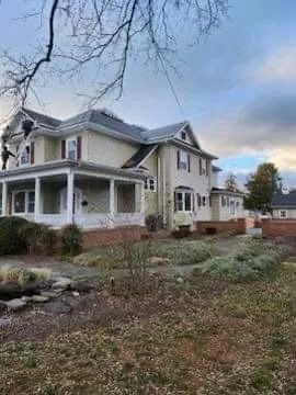 Large two-story house with wrap-around porch, light yellow siding, and brick foundation; cloudy sky.