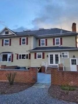 Two-story beige house with red shutters, brick wall, and walkway. Cloudy sky in the background.