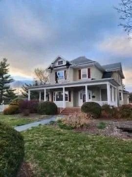 Two-story house with wrap-around porch, yellow siding, red shutters, and manicured lawn. Cloudy sky.