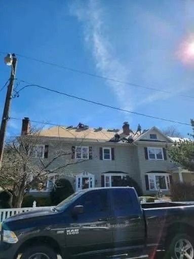 Two-story house with roof damage under a bright blue sky. A truck is parked in front.