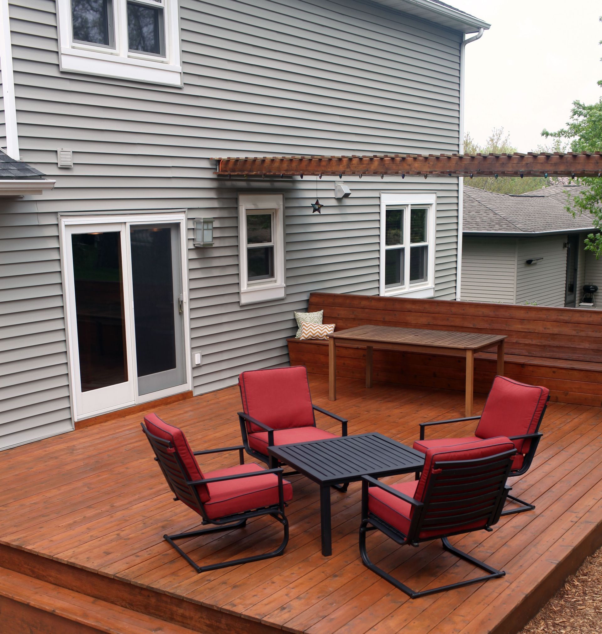 Wooden deck with red chairs and table, connected to a gray house with a pergola.