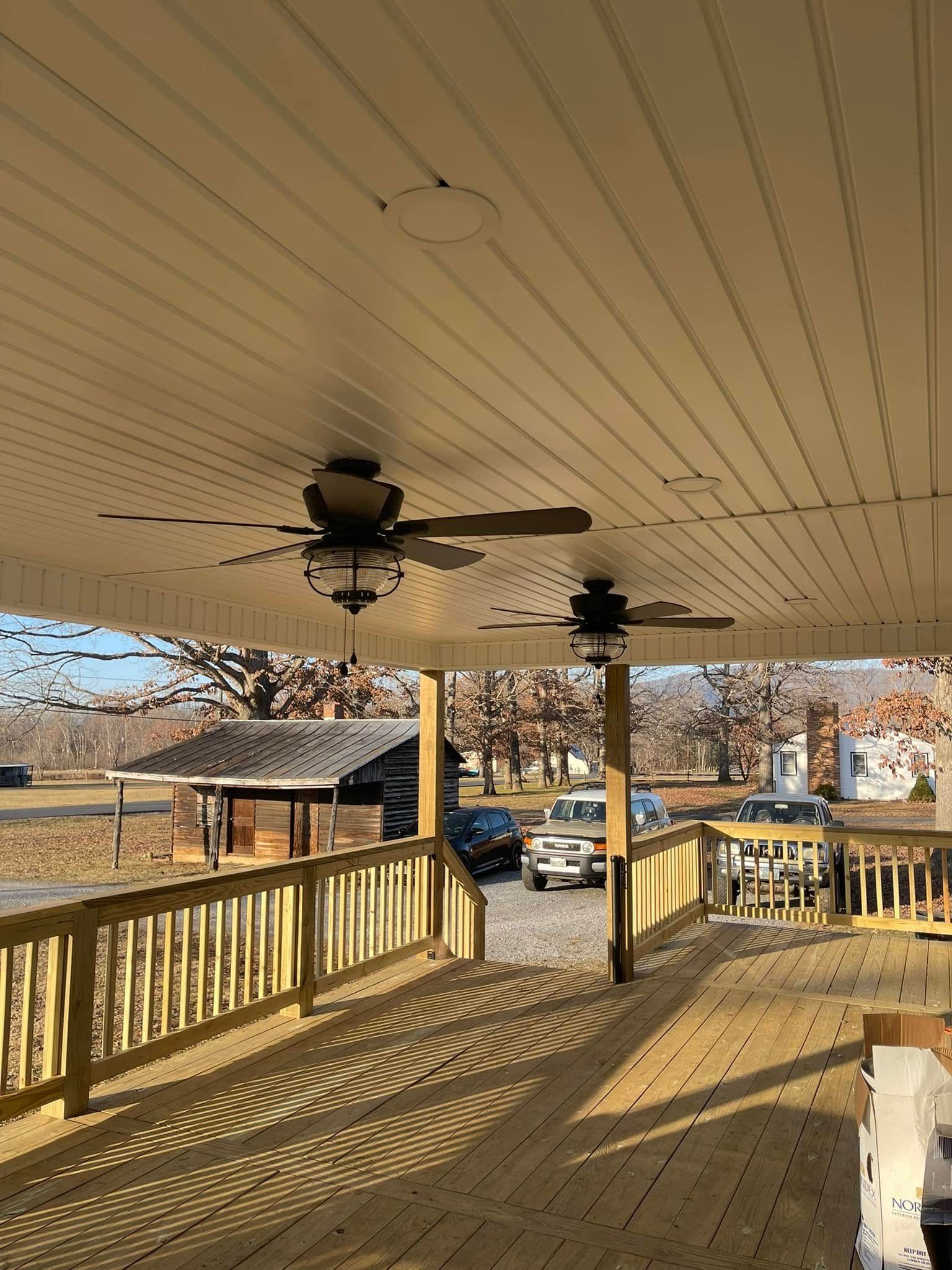 Covered wooden porch with ceiling fans, railings, and a view of parked cars and trees beyond