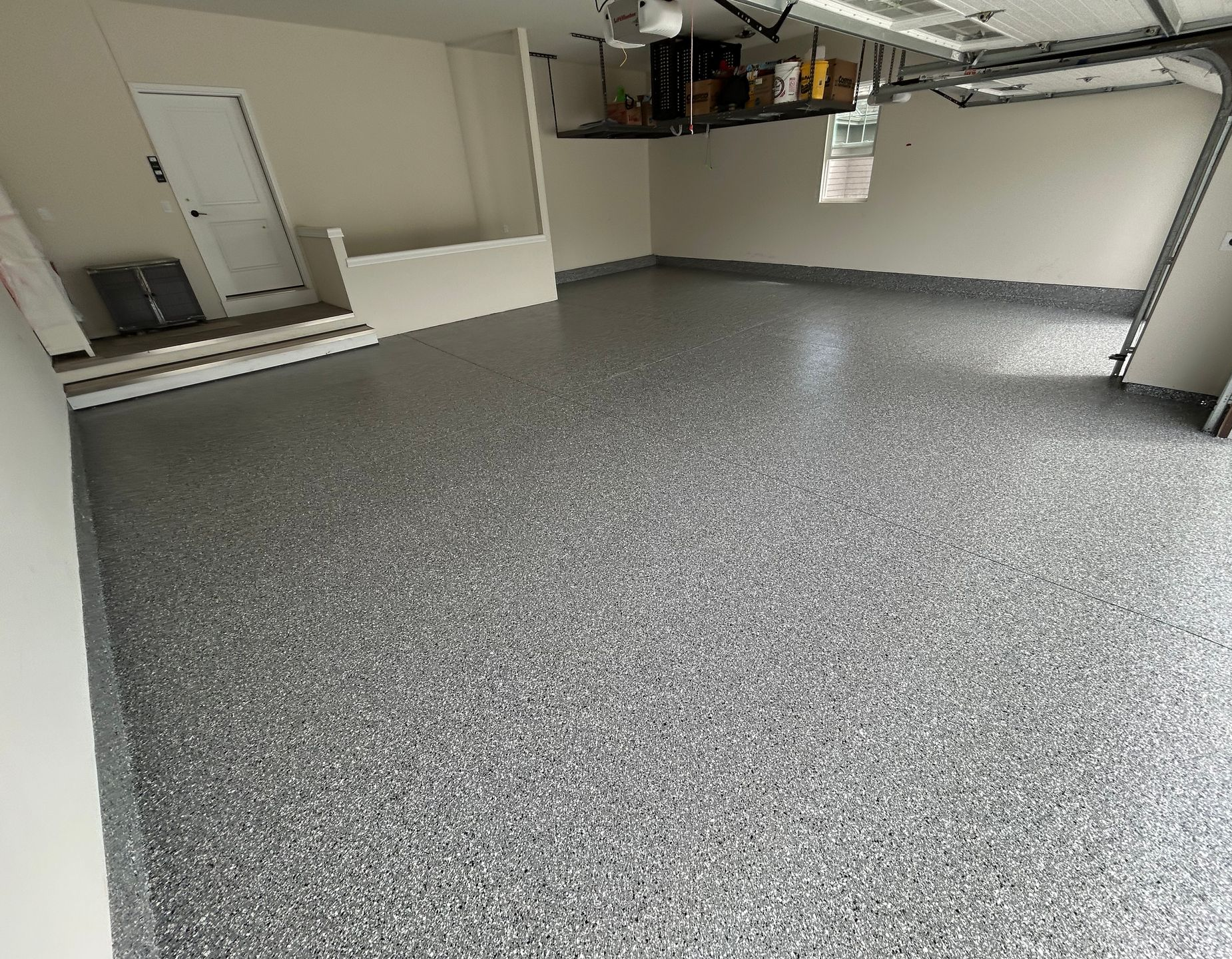 Empty garage with gray speckled floor, beige walls, and exposed ceiling pipes and storage shelves