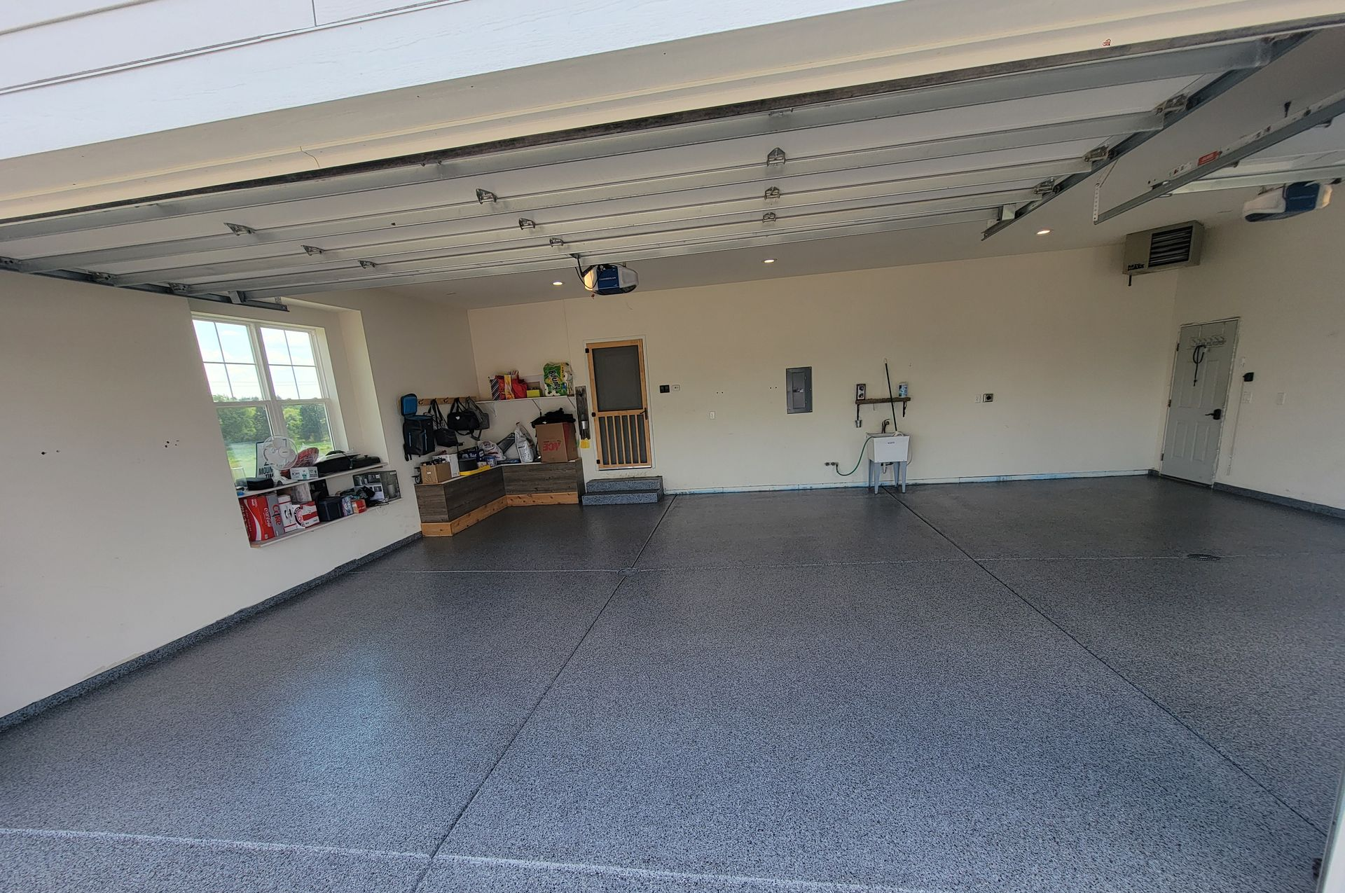 Empty gray-floored garage with an open door, storage shelves, and a small workbench along the back wall