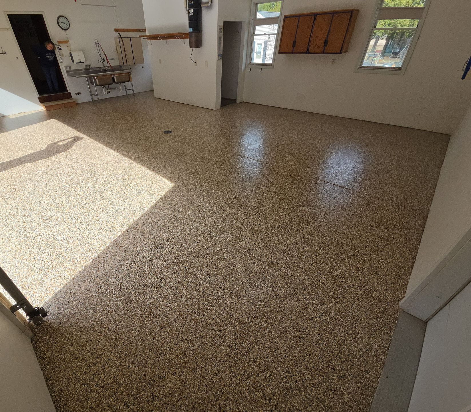 Empty sunlit room with speckled floor, white walls, windows, and kitchen cabinets in back
