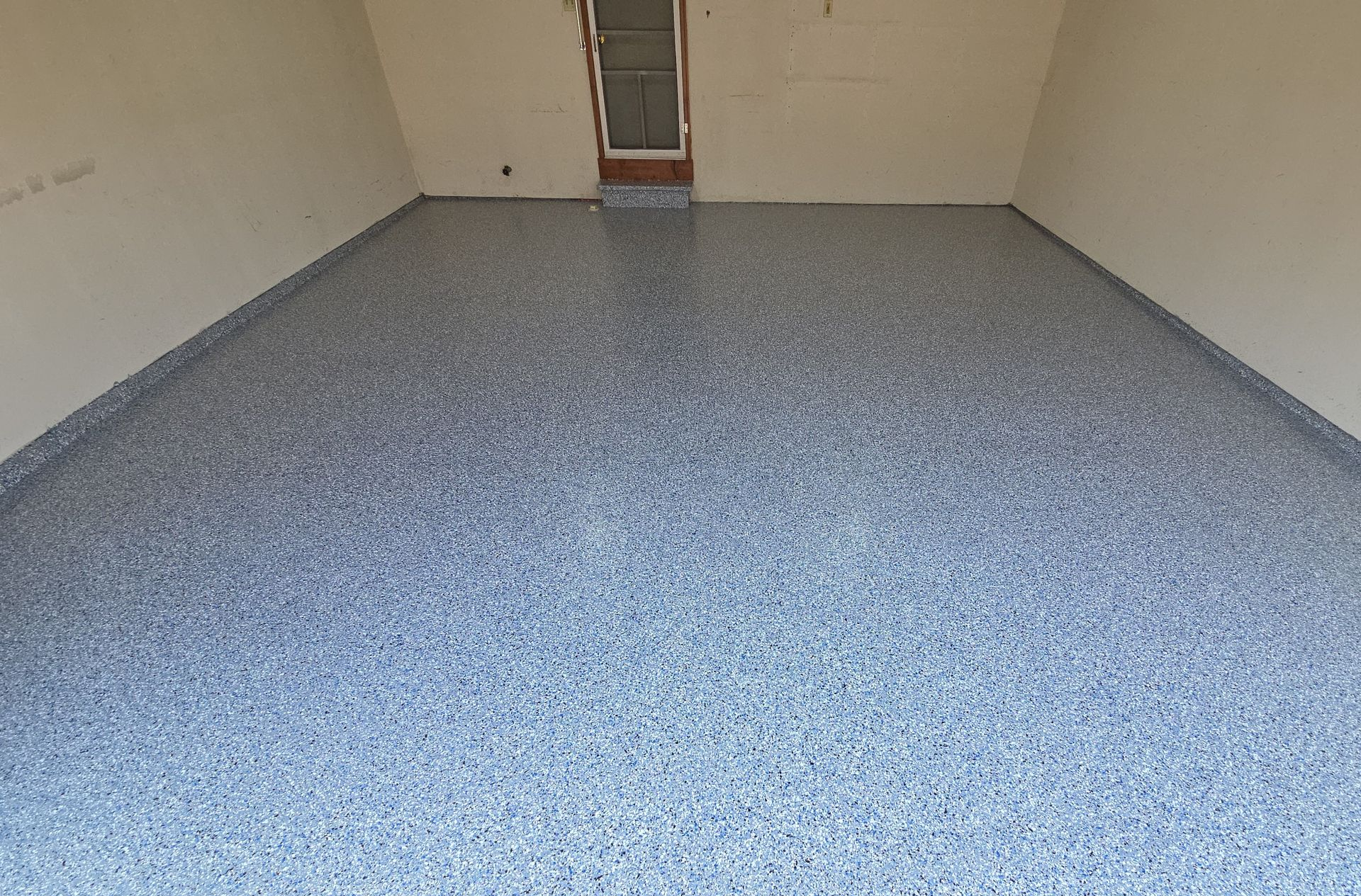 Empty garage with speckled gray floor and beige walls, viewed toward a closed door at the back.