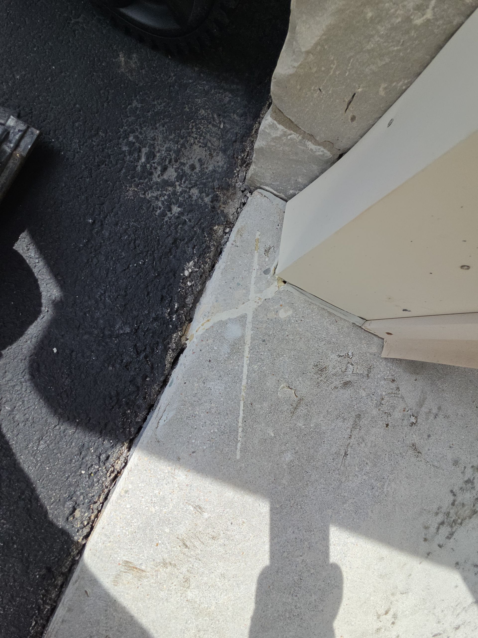 Corner of a walkway with black asphalt beside a light concrete curb and wall in sunlight