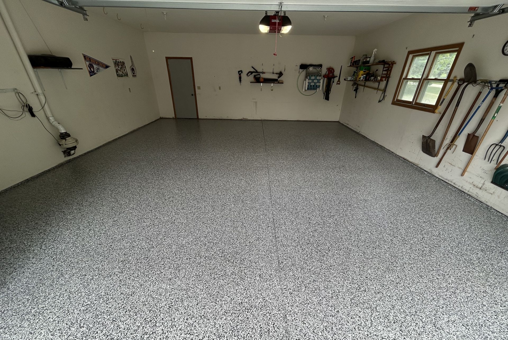 Empty garage with speckled gray floor, white walls, tools hanging on the right wall, and a window on the far side