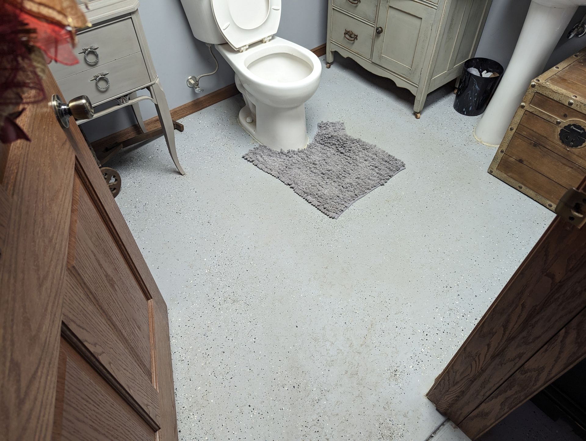 Small bathroom with white toilet, gray floor, rug, cabinet, and wooden furniture.