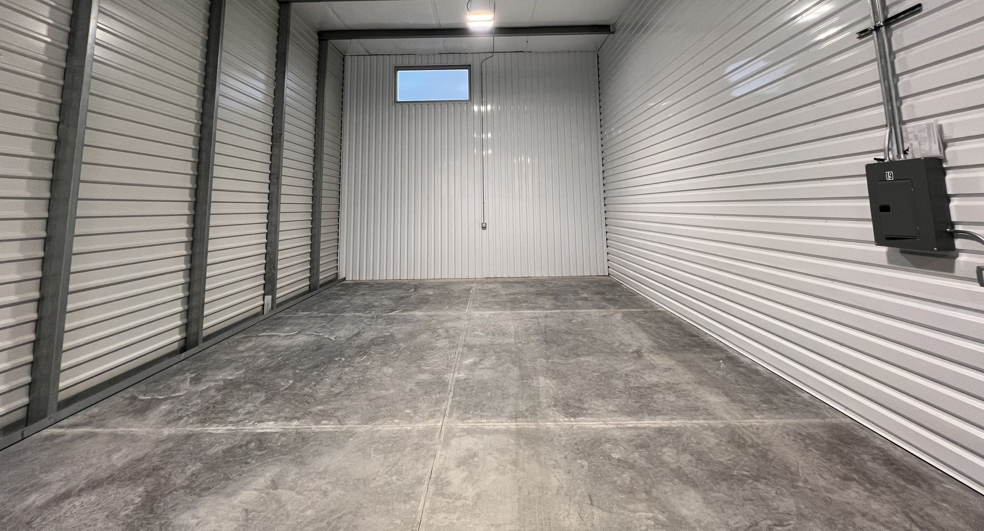 Empty metal storage unit with gray floor and white corrugated walls, viewed from the entrance.