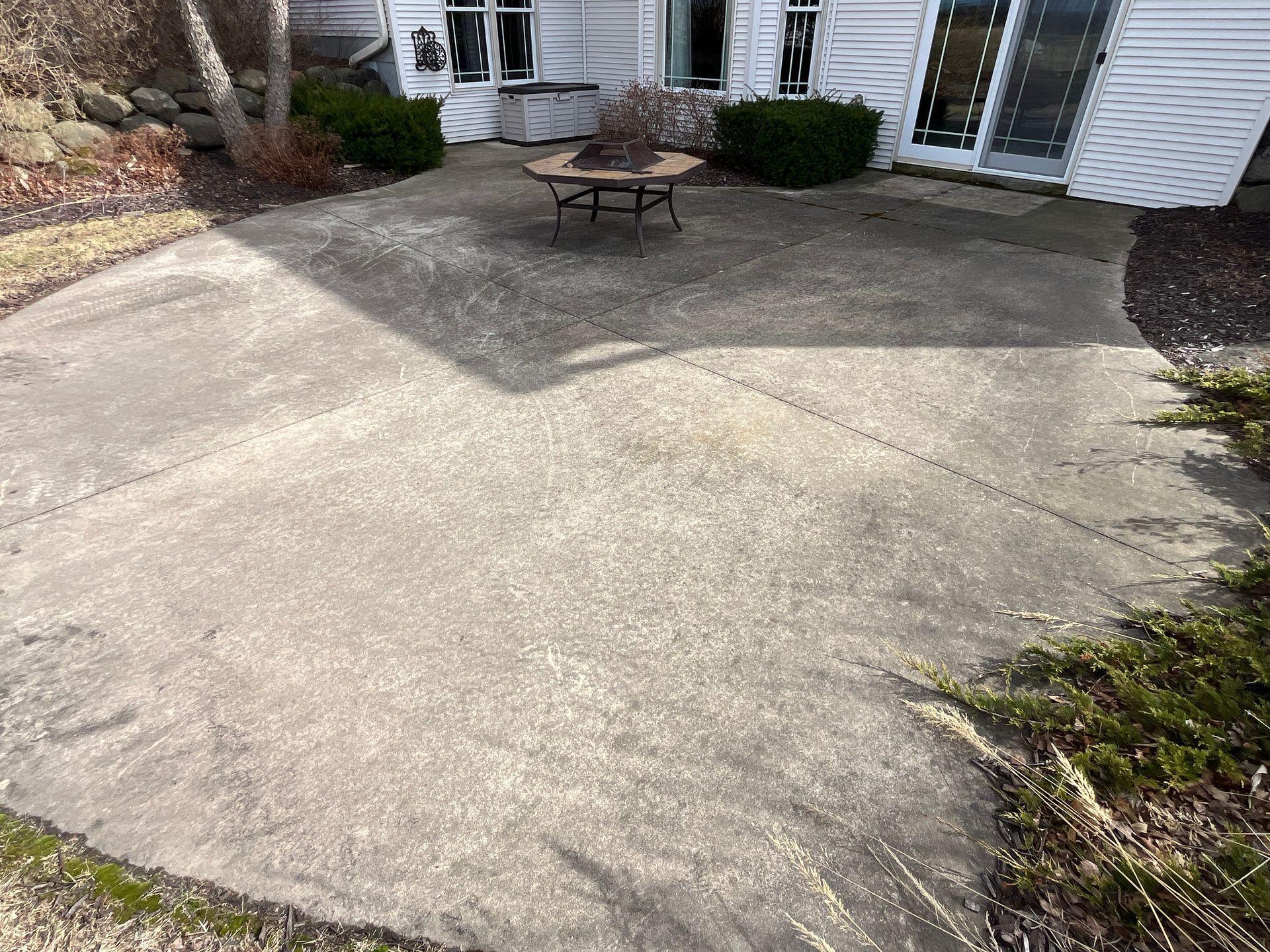 Concrete patio area with a small table and chairs beside a white house and bushes