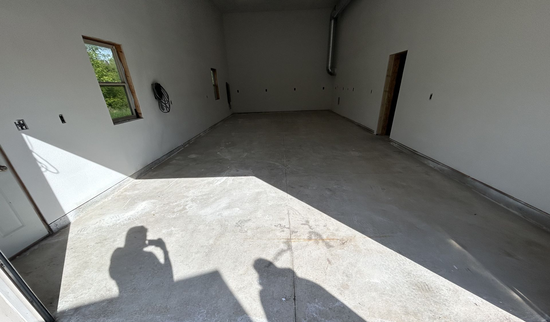 Empty unfinished room with concrete floor, white walls, windows, and strong sunlight casting shadows.