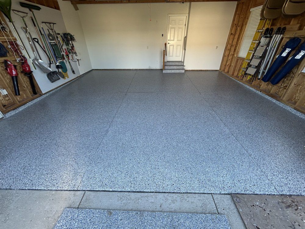 Empty garage with gray epoxy floor, wall-mounted tools, and a staircase at the back.