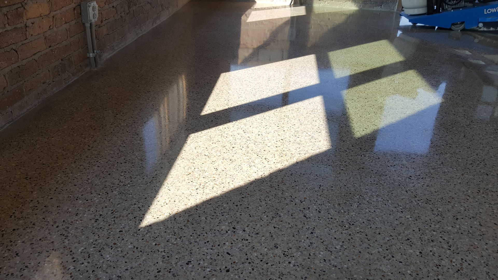 Sunlit terrazzo floor with sharp window shadows and a brick wall along the left side