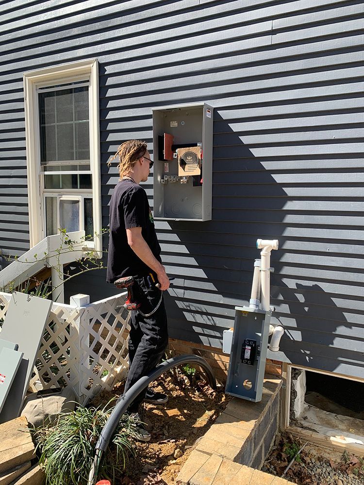 A person works on electrical box attached to grey house siding, another box lies below.