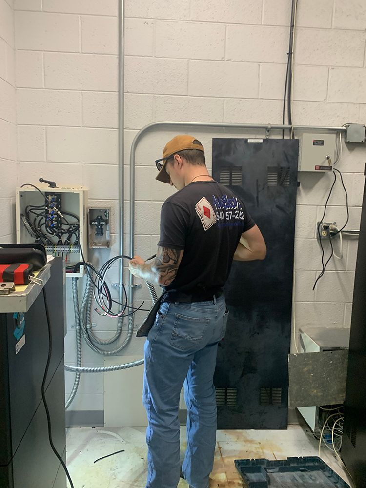 Man in a baseball cap and safety glasses, working with wires near electrical panels in a utility room.