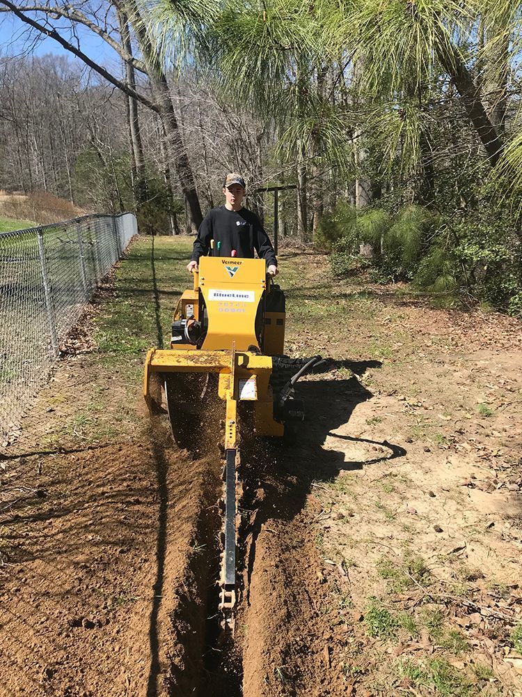 Man operating a yellow trencher, digging a trench along a fence in a wooded area.