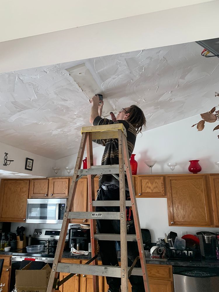 Person on a ladder repairing a ceiling in a kitchen. Wearing a patterned shirt, working on a damaged area.