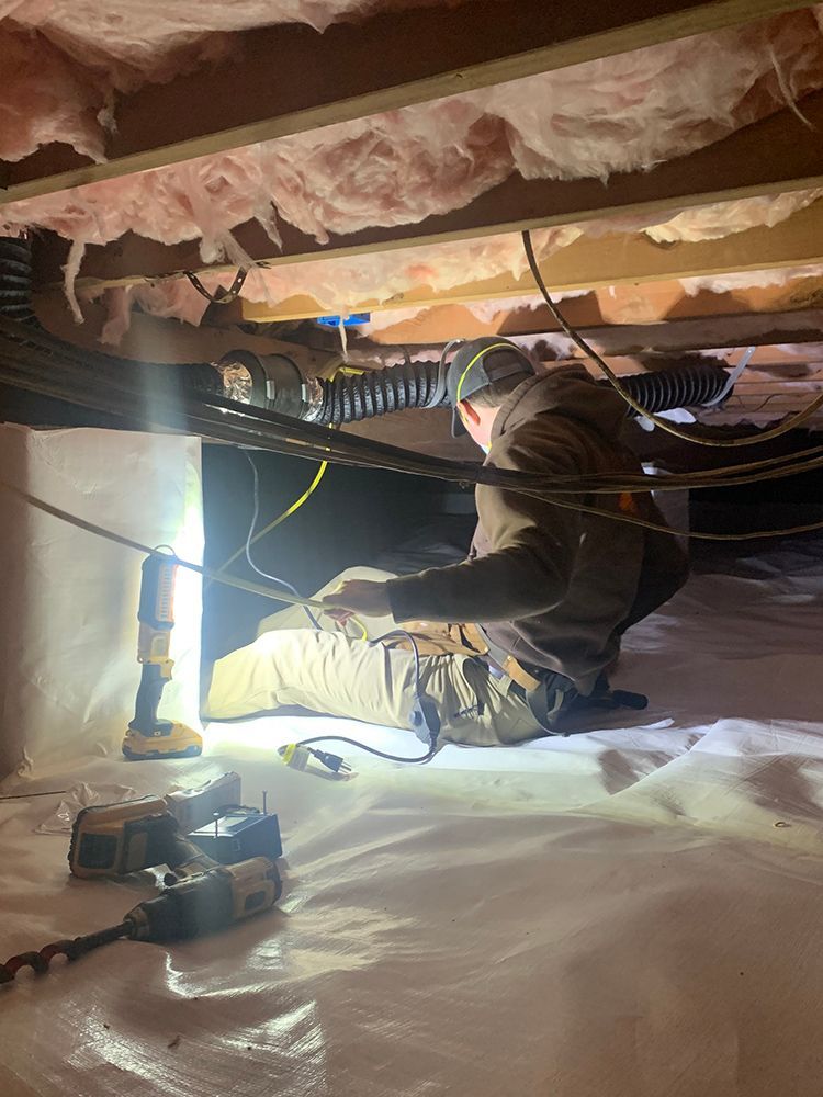 A worker crouches in a crawl space, inspecting wiring with a headlamp, tools and a flashlight visible.