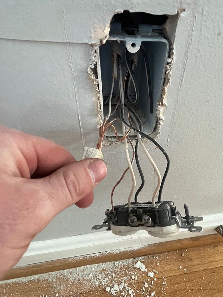 Hand holding wire connector near electrical outlet with exposed wires in wall.