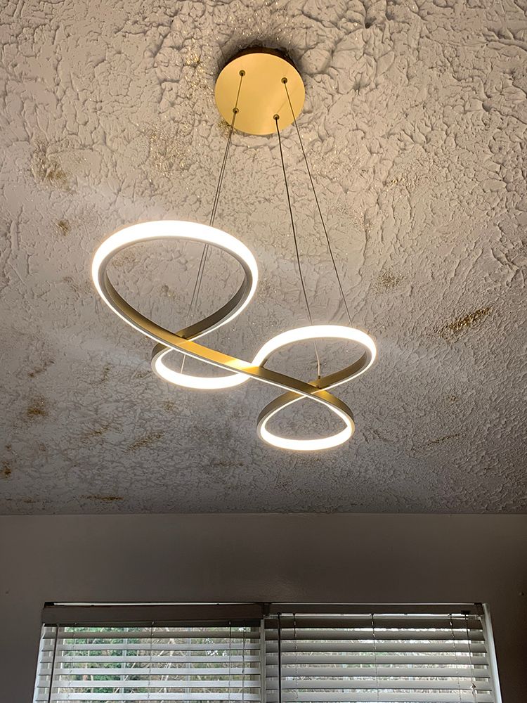 Gold ring-shaped LED chandelier against a stained ceiling, above a window with blinds.